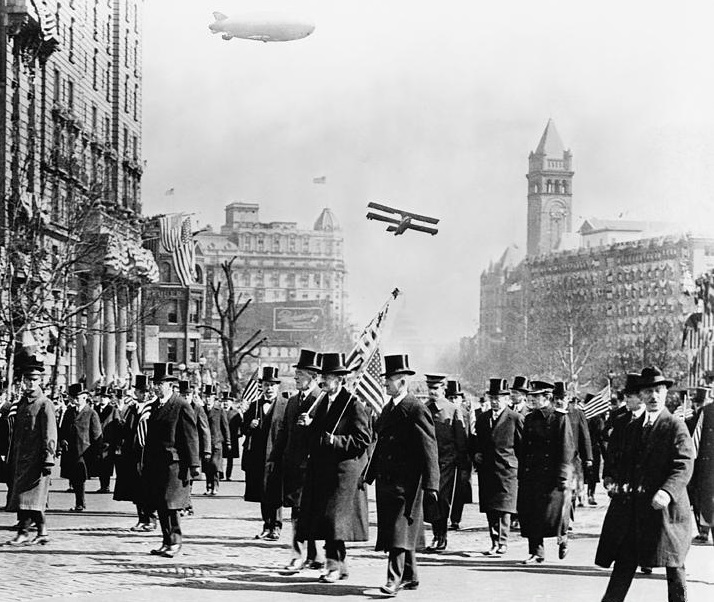 A group of people in a parade. A blimp and plane fly by.